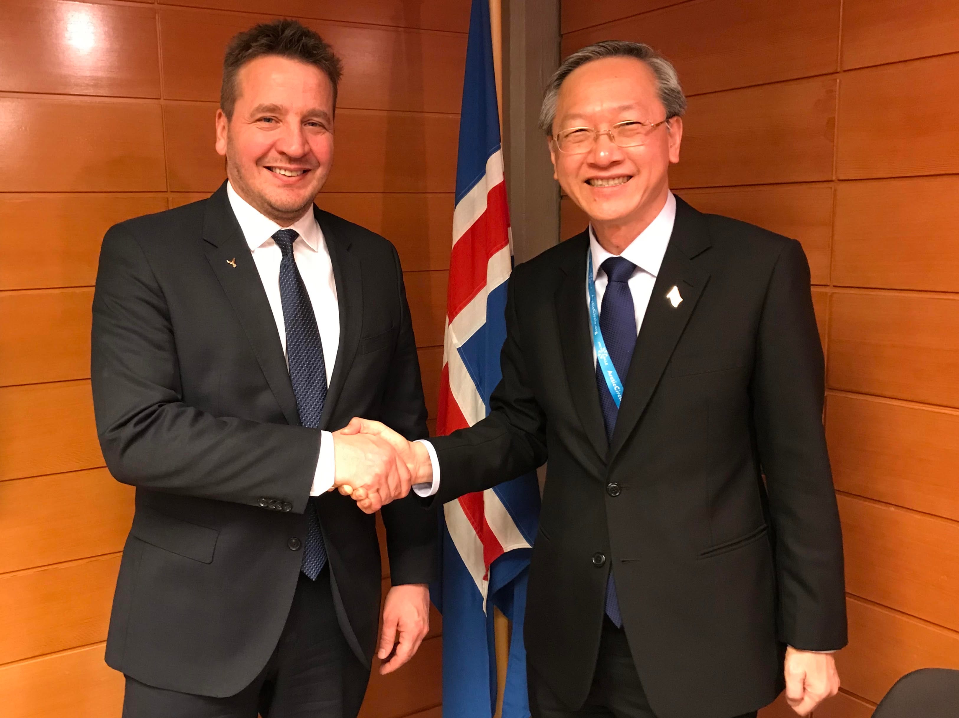 Two men in suits shake hands, Iceland flag behind them.